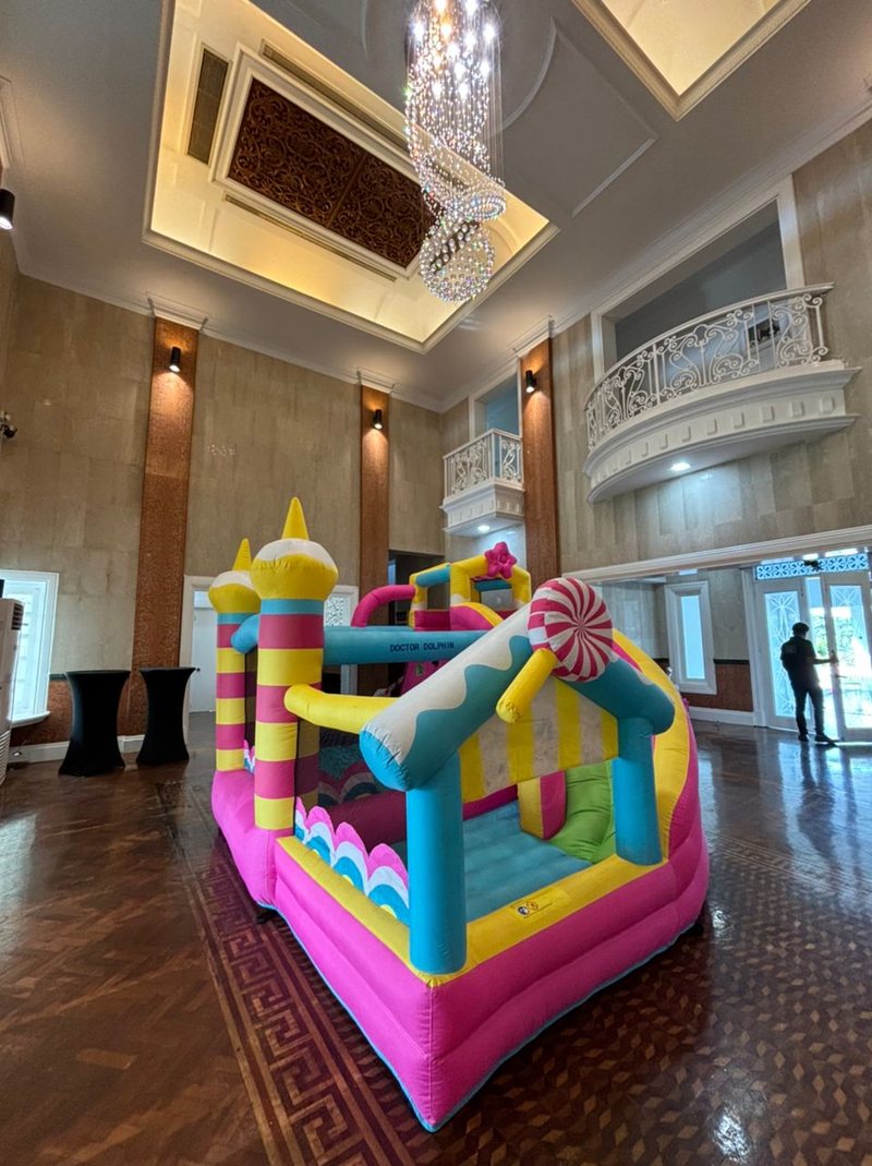 Pink and yellow candy-themed bouncy castle standing on a parquet floor in a high-ceilinged ballroom with a chandelier