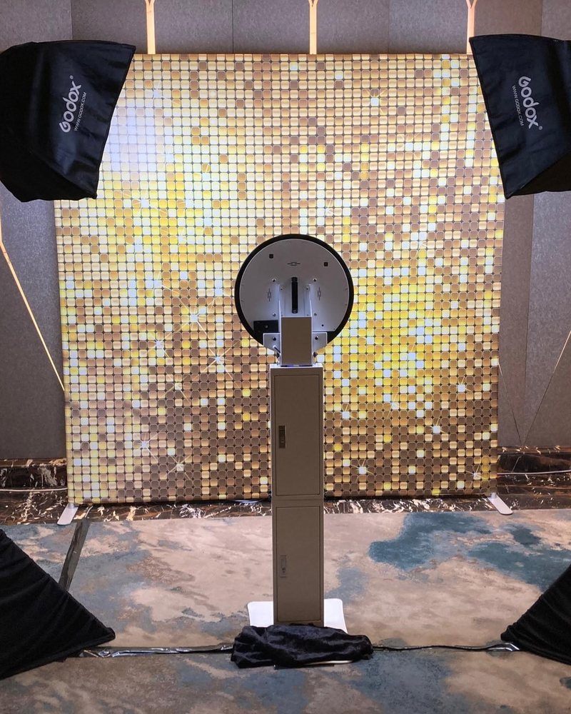 Digital photo booth on a slim white stand in front of a gold sequin backdrop, lit by two softboxes in a ballroom