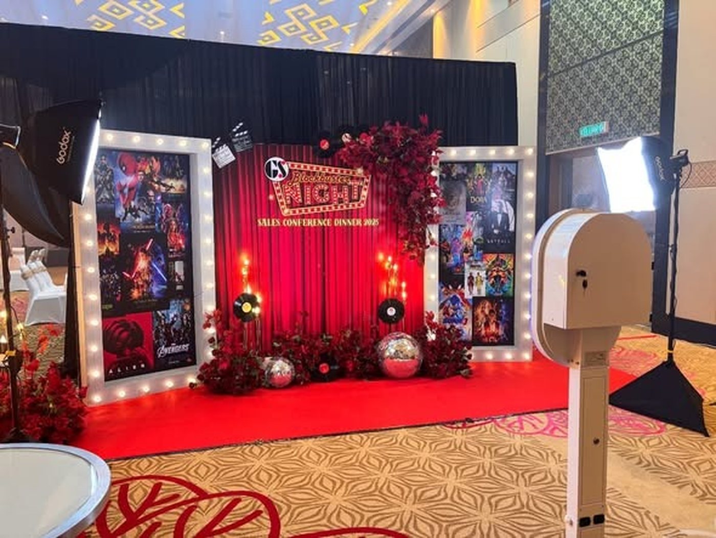 DSLR photo booth and studio lights set up beside a themed backdrop and red carpet in a hotel ballroom before guests arrive