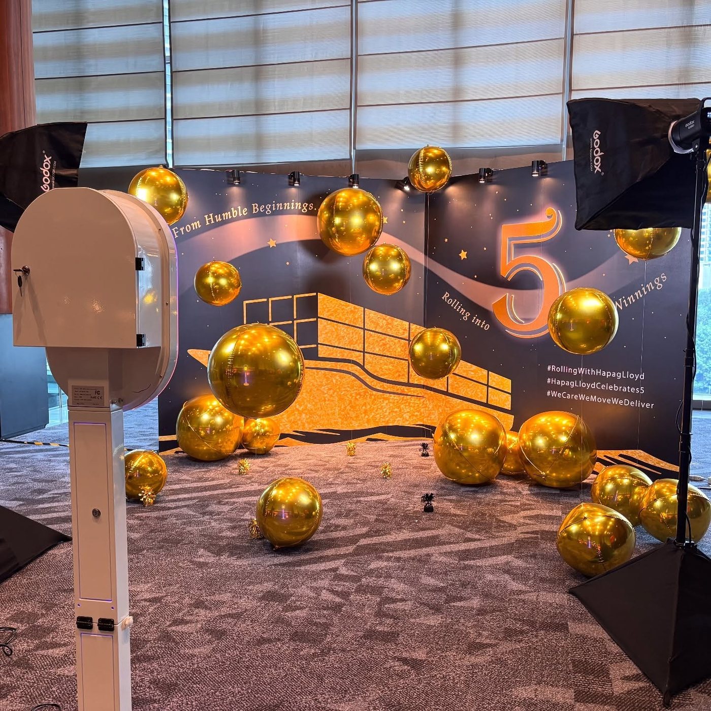 Photo booth unit and softbox lights positioned in front of a corporate anniversary backdrop dressed with gold balloons